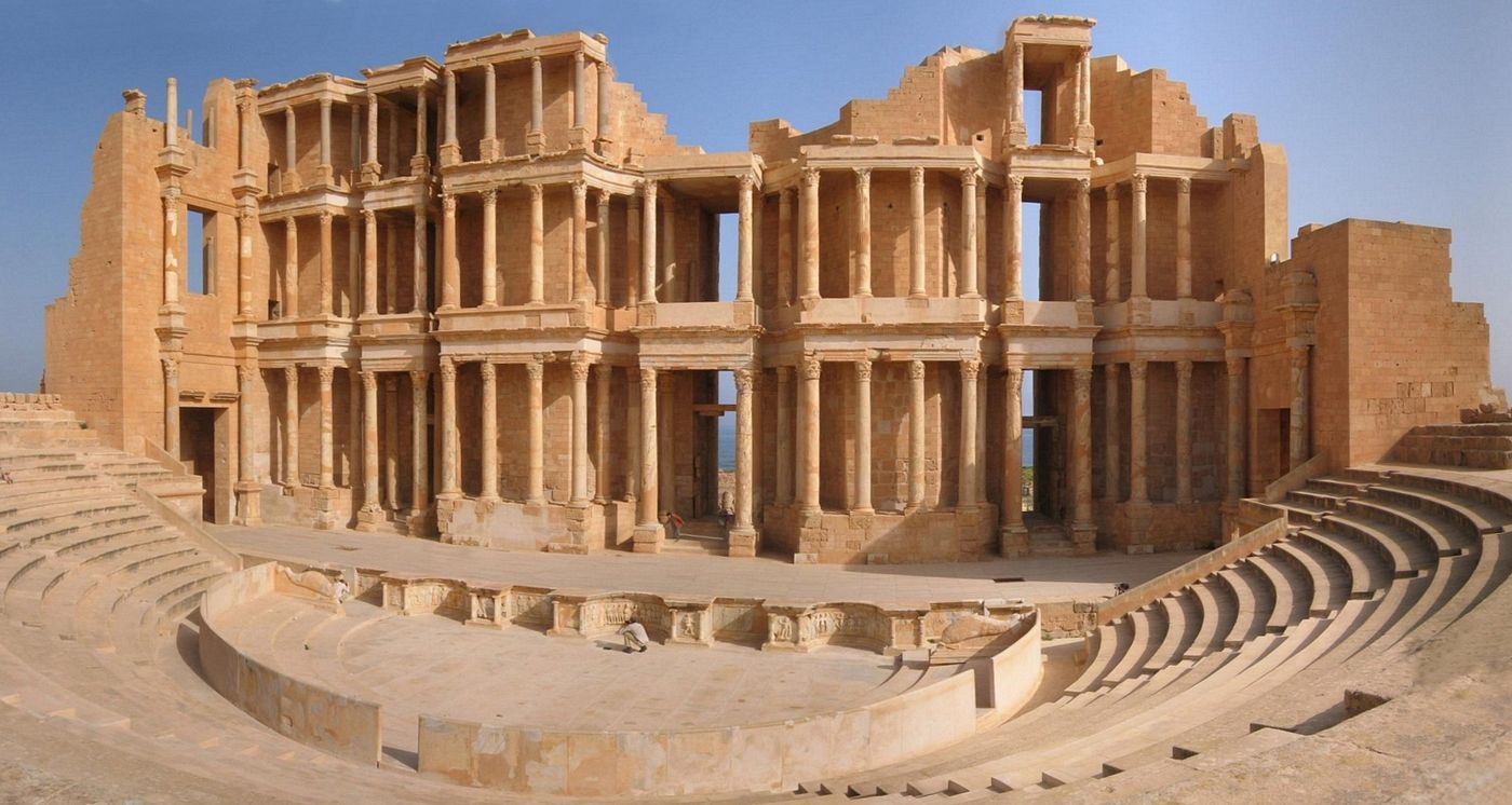 The tiered stone seating and reconstructed columned stage building of the Roman theatre at Sabratha, under a blue sky.