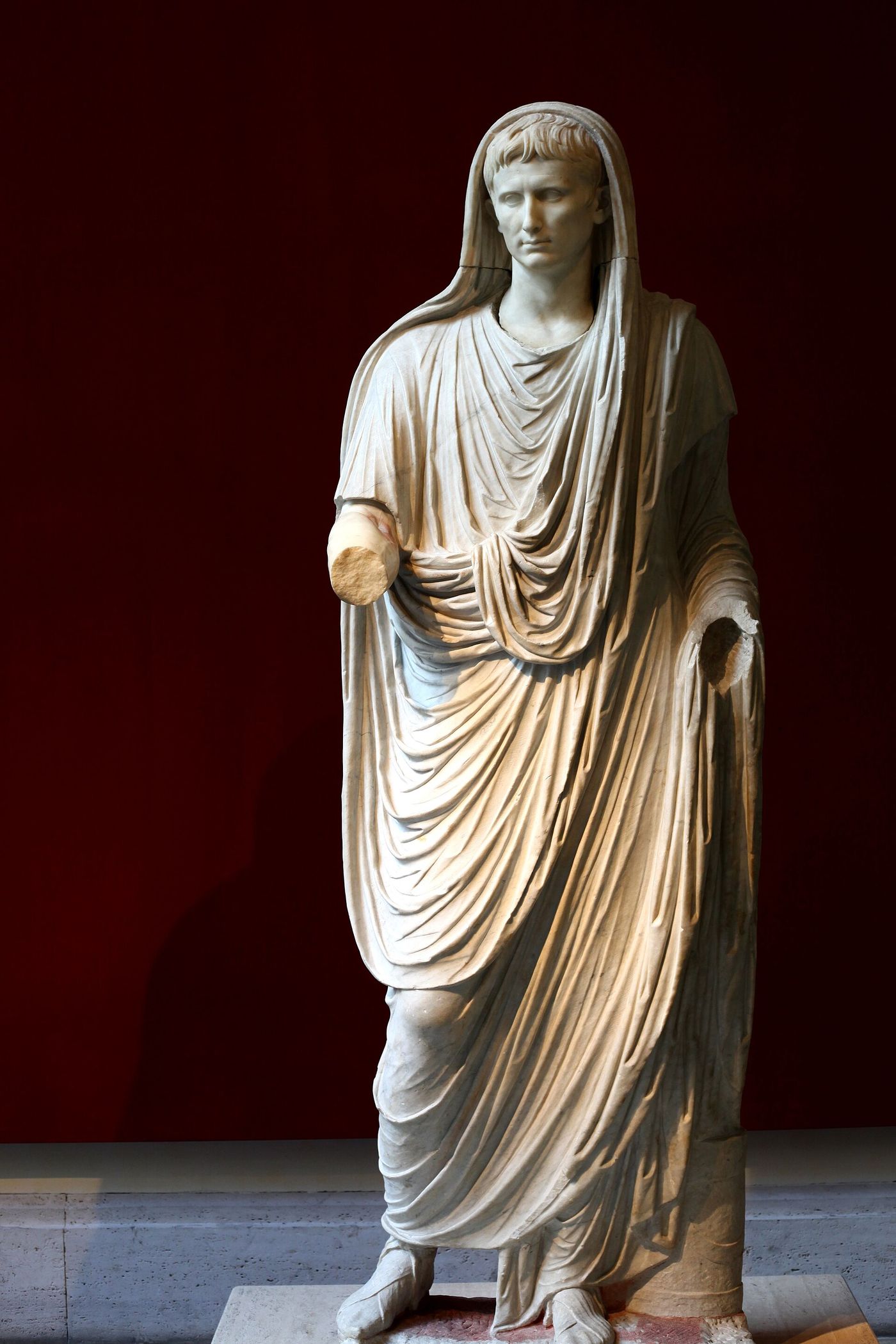 A marble statue of a man in a heavy toga with a fold of the cloth drawn up over his head, both forearms broken away, standing against a dark red wall.