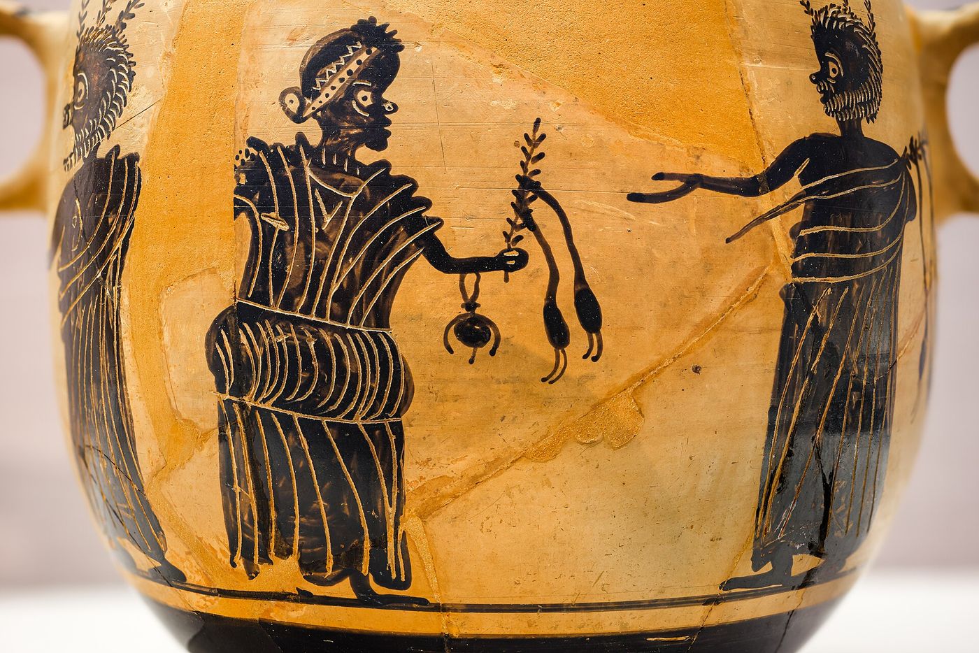 Detail of a Greek black-figure vase: a seated figure with an exaggerated grotesque face and a patterned headband holds out a small jug and a leafy sprig towards a standing figure with wild spiky hair.