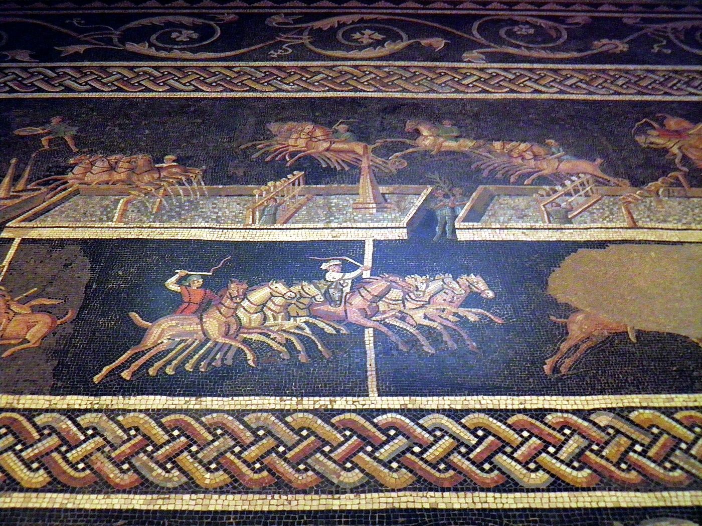 A Roman mosaic showing four-horse chariots racing around a central barrier, with charioteers in coloured tunics driving them.
