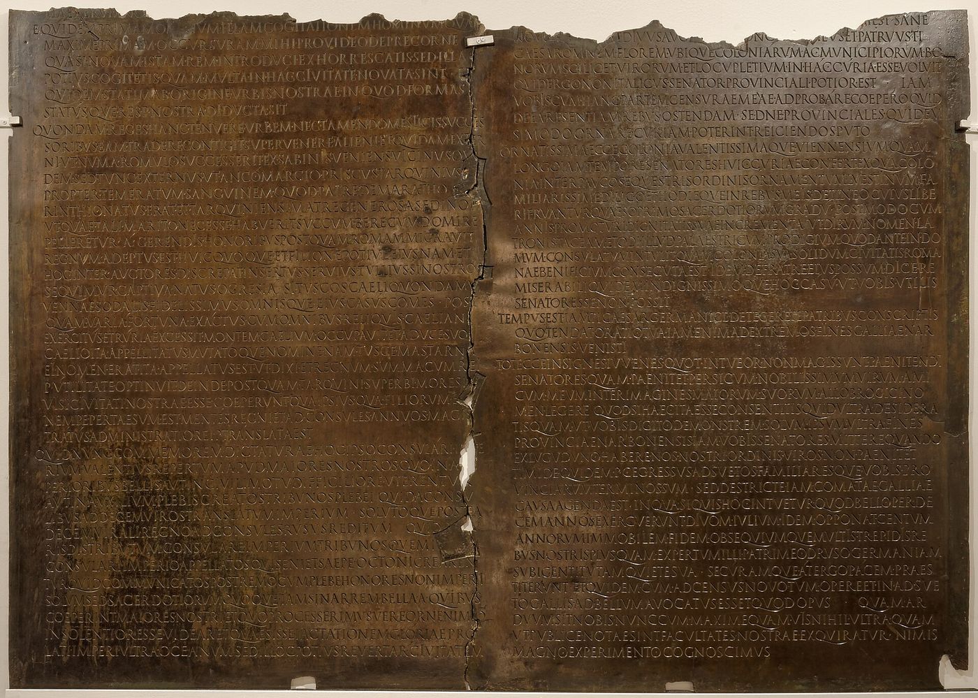 A large bronze plaque engraved with two dense columns of Latin capitals, split by a ragged vertical break down the centre and broken away along the top edge.