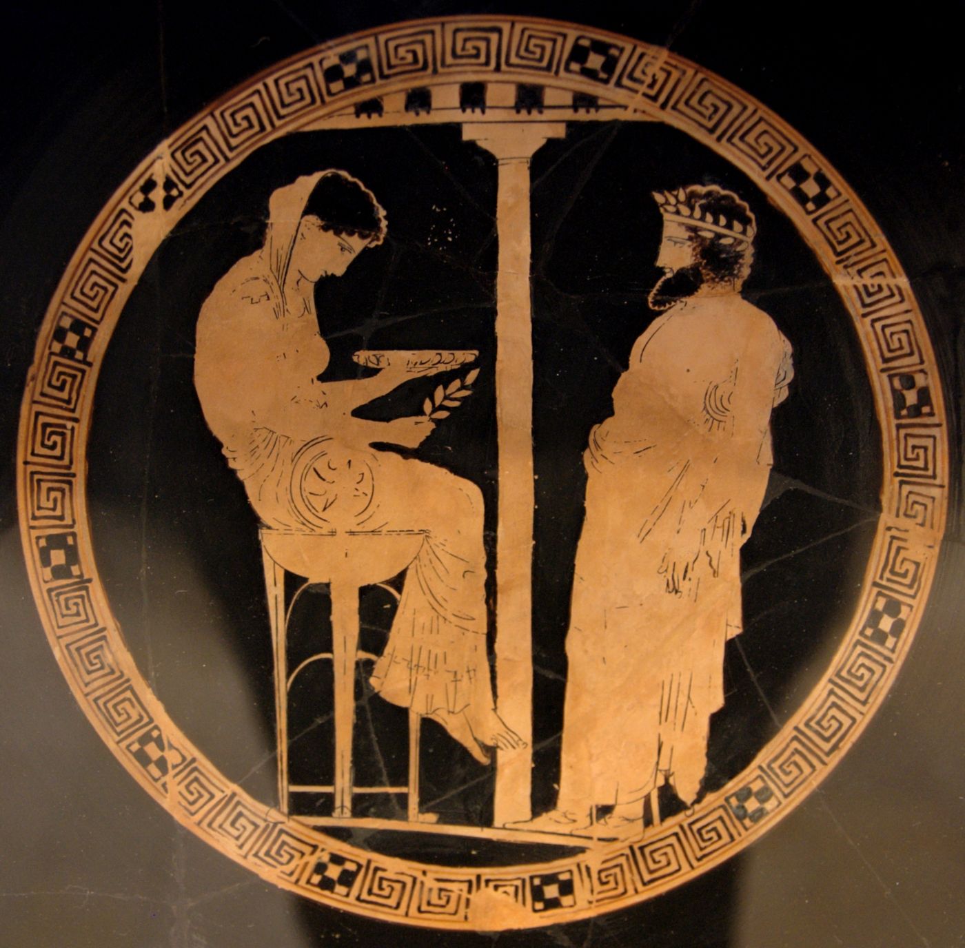 The interior of a Greek red-figure cup: a woman sits on a tall tripod, head bowed over a shallow bowl held in one hand and a laurel sprig in the other, facing a bearded man in a wreath who stands wrapped in his cloak.
