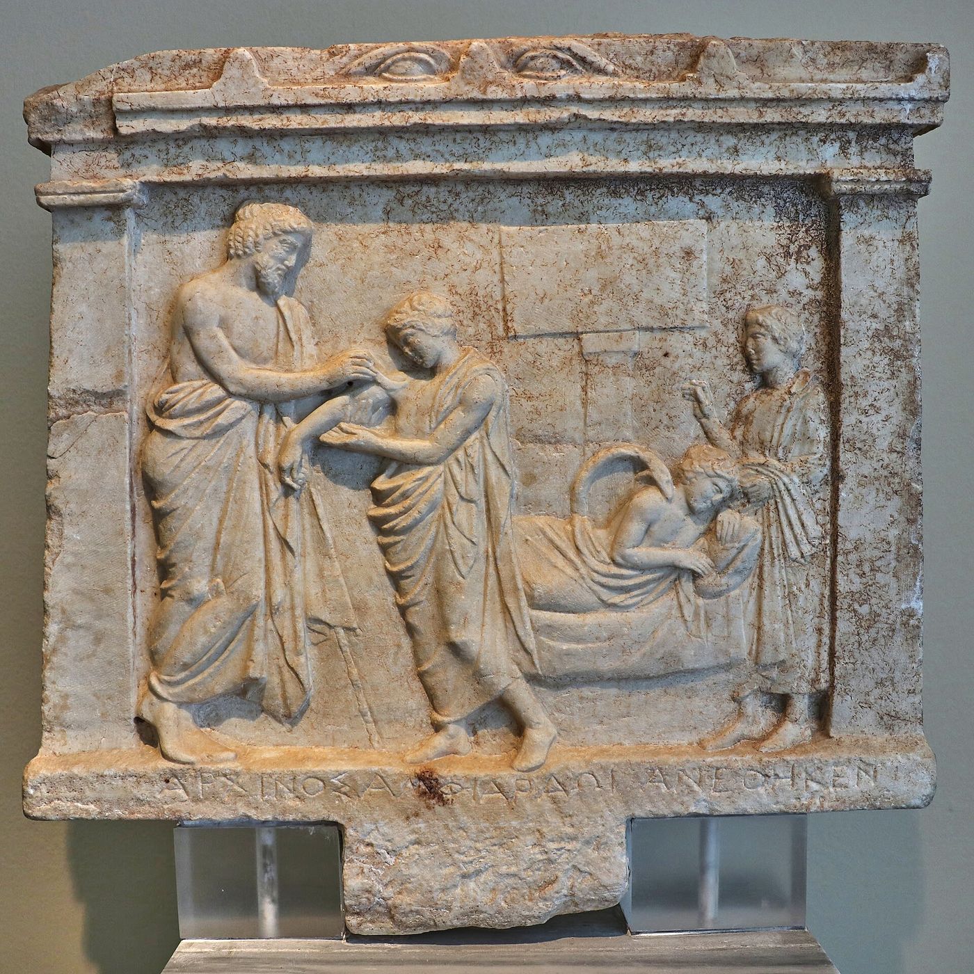 A marble votive relief: a bearded god treats the bare shoulder of a standing young man, while to the right the same young man lies asleep on a couch with a large snake stretched over his shoulder.