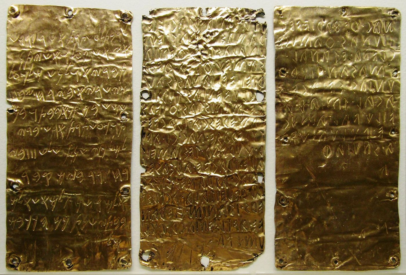 Three thin rectangular sheets of gold displayed side by side, each crumpled slightly and covered in rows of incised ancient lettering, with small nail holes around their edges.