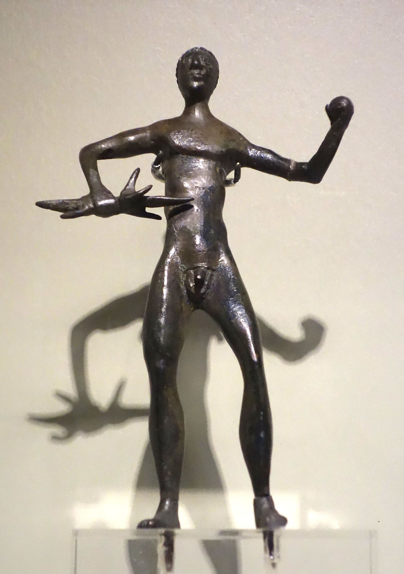 A slender nude bronze statuette of a standing male figure, one arm extended holding a forked thunderbolt, the other raised holding a small round object.