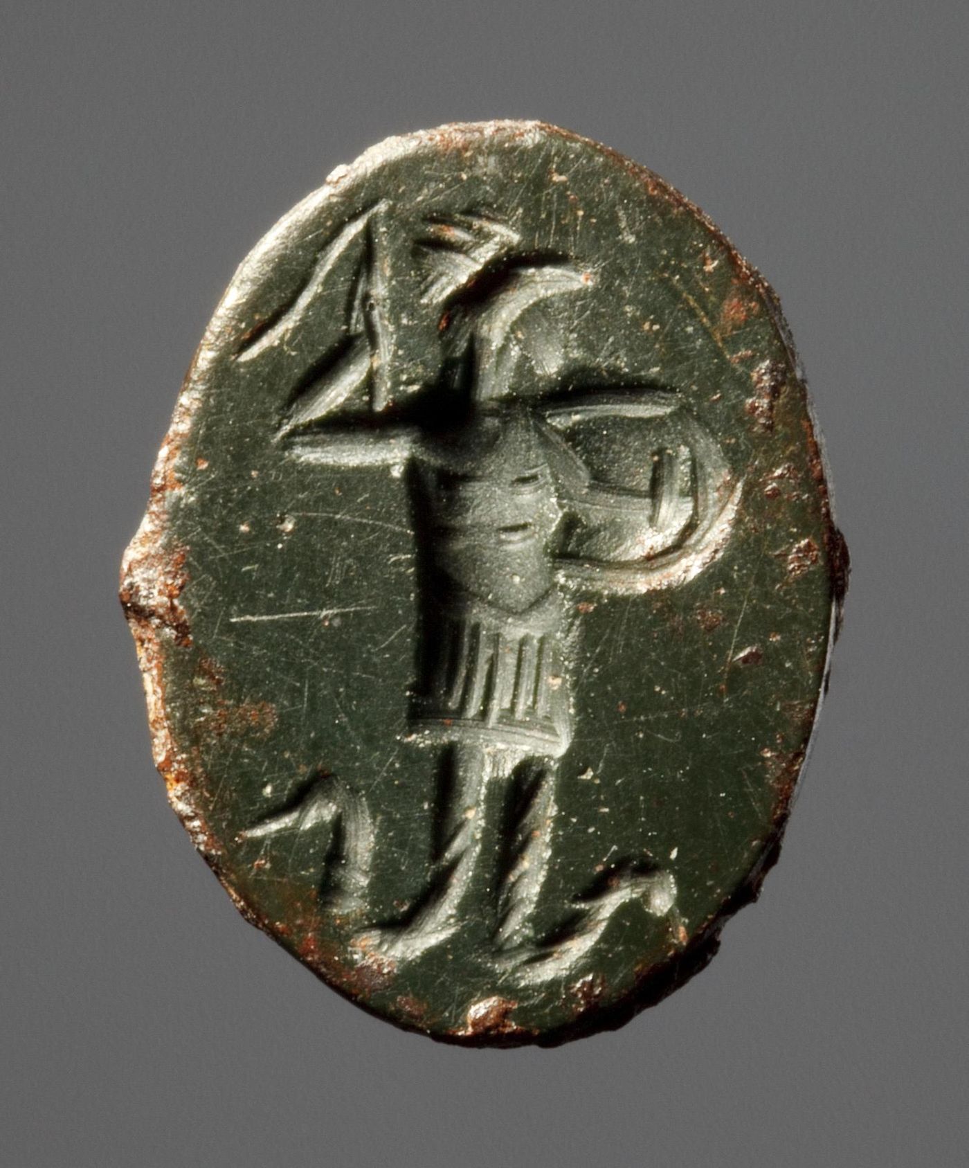 A carved oval gemstone showing a figure with a rooster's head, holding a shield and whip, with serpents in place of legs.