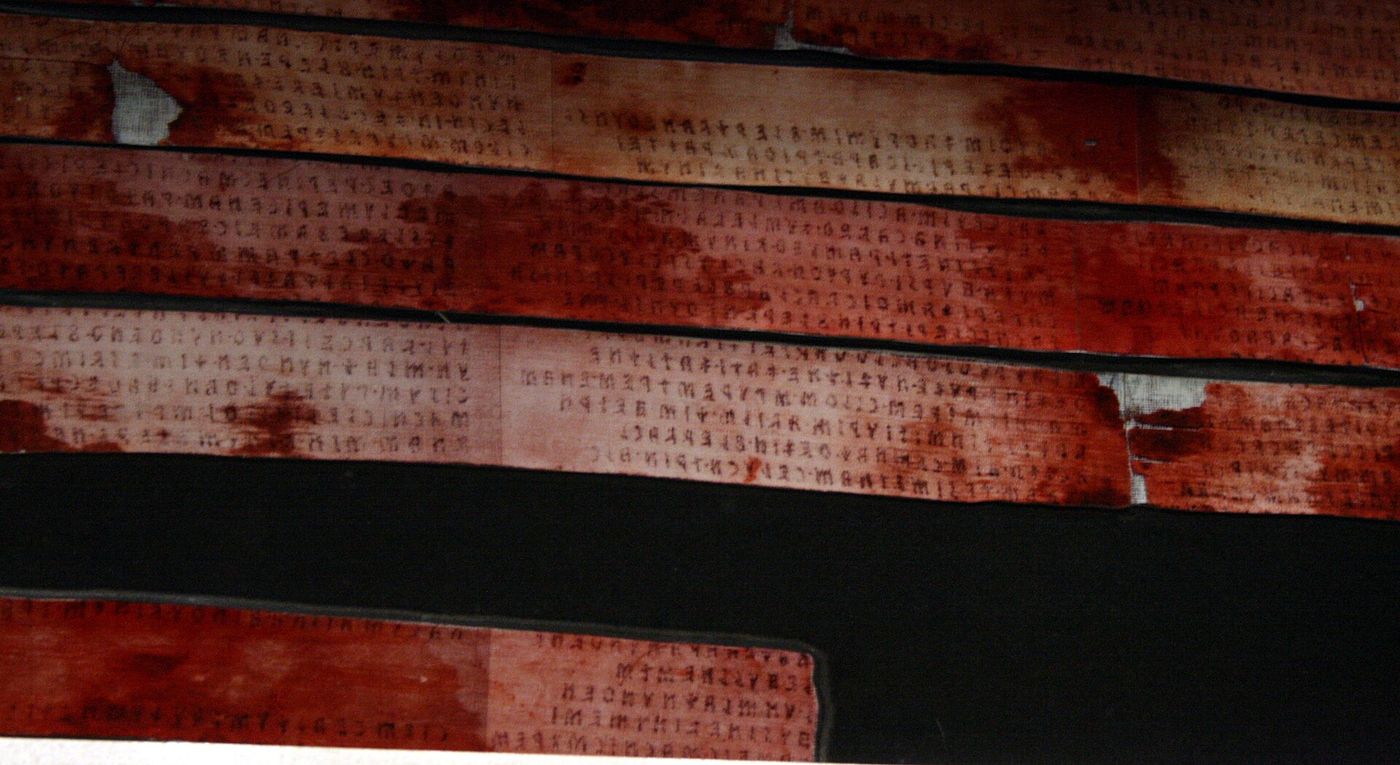 Long horizontal strips of ancient linen, stained deep red-brown, each carrying rows of small dark Etruscan letters, mounted against a black backing.