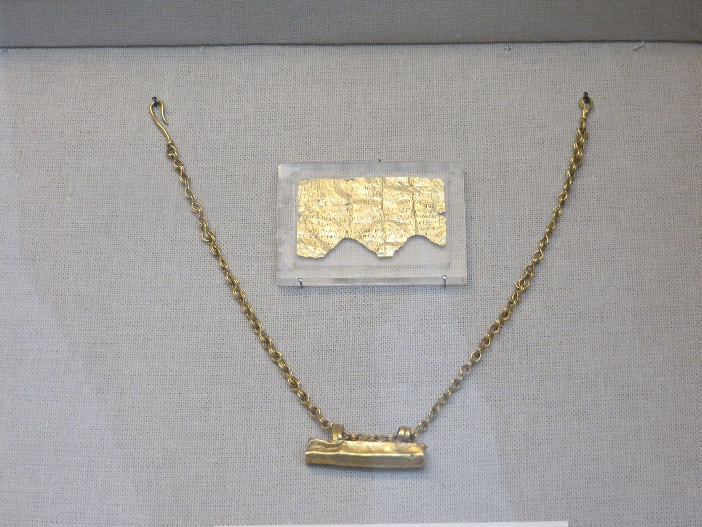 A small rectangular sheet of gold leaf covered in tiny incised Greek letters, displayed in a museum case.
