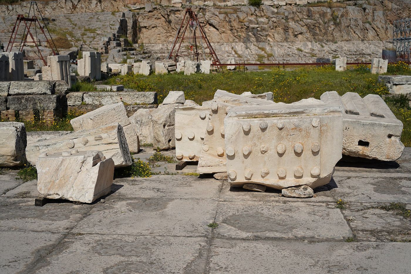 Large fallen marble blocks, their faces carved with rows of projecting knobs, lying on stone paving at Eleusis with a rock-cut terrace rising behind them.